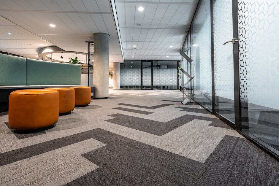 A modern office lobby featuring a black rectangular rug on a polished black and white patterned tile floor. Four blue upholstered armchairs with wooden legs are arranged around a small round black cof