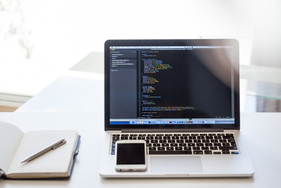 A close-up view of a modern workspace showing a silver MacBook laptop with a screen displaying a code editor filled with colorful programming code, placed on a clean white desk. To the left of the lap